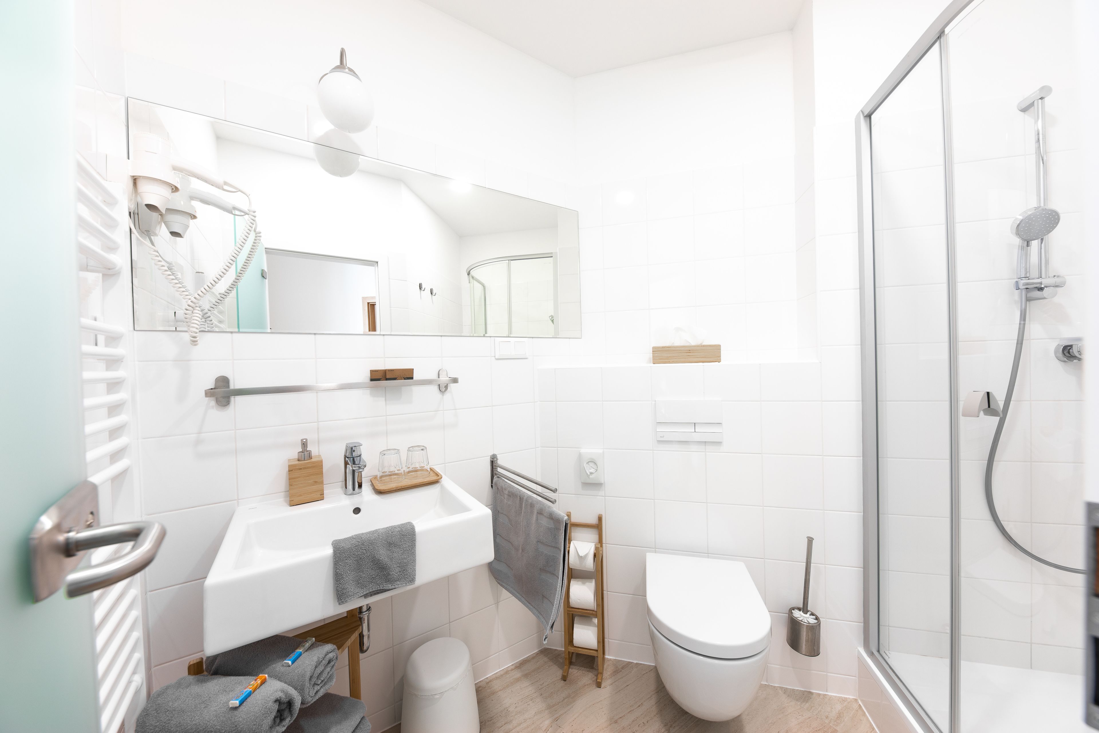 Un baño moderno con un lavamanos, espejo y retrete blancos, una zona de ducha y un perchero para toallas.