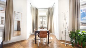 A dining area with a wooden herringbone floor, a table with chairs, a hanging light fixture, and a view of buildings through a window with curtains. - Hotel and Apartments Jacob (Brno)