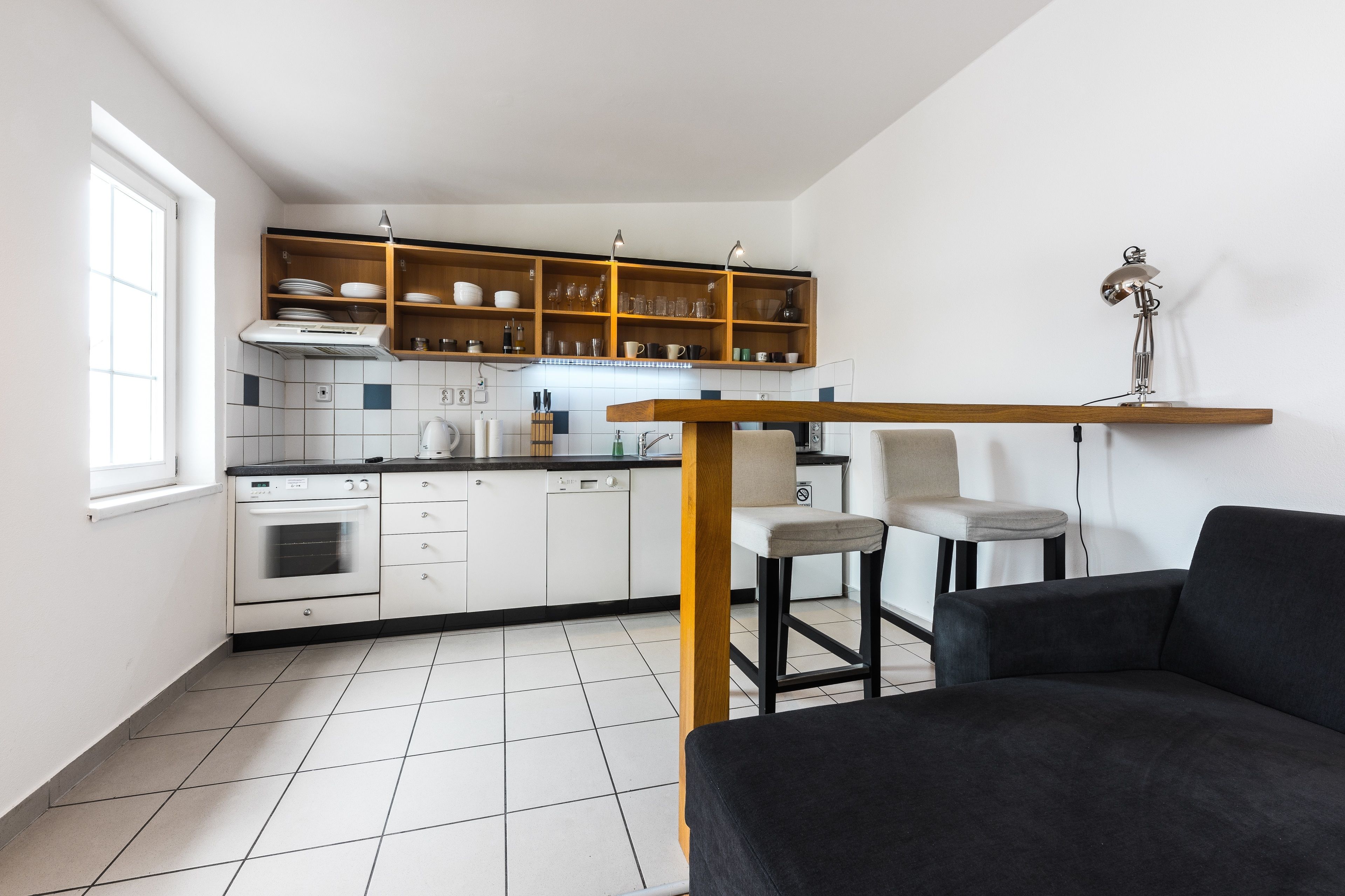 A compact kitchen with white cabinets, a wooden countertop, and a built-in shelf above the sink. There is a black sofa and two chairs in the living area.