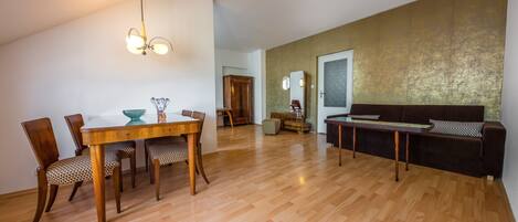 A spacious living area with a wooden floor, a dining table with chairs, a sofa, and a ceiling light fixture.