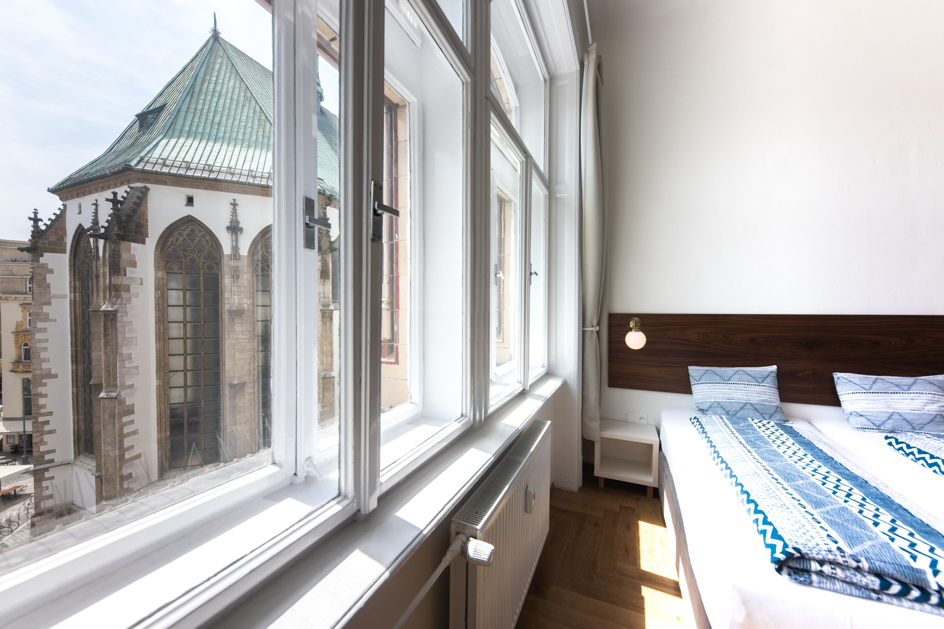 Un dormitorio con un ventanal grande, una cama con ropa de cama estampada y vistas a un edificio histórico con techo verde.