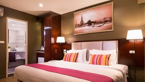 A hotel room with a large bed, a nightstand, a desk with a lamp, a bathroom with a sink and mirror, and a wall-mounted picture of a cityscape. - Ratchada Point Hotel (Bangkok)