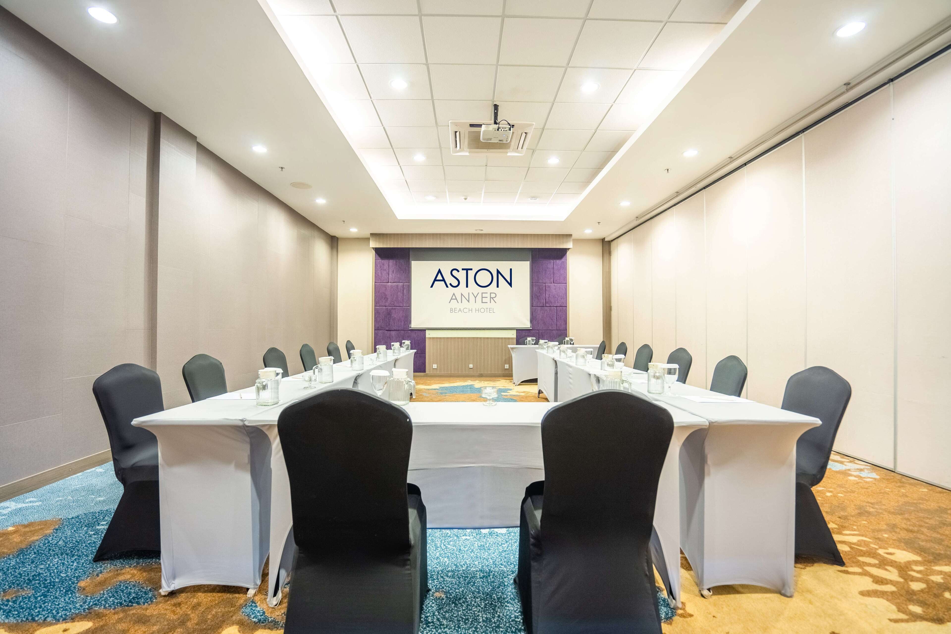 Un salón de conferencias con una larga mesa, sillas negras y una pantalla que muestra 'ASTON ANYER BEACH HOTEL'.