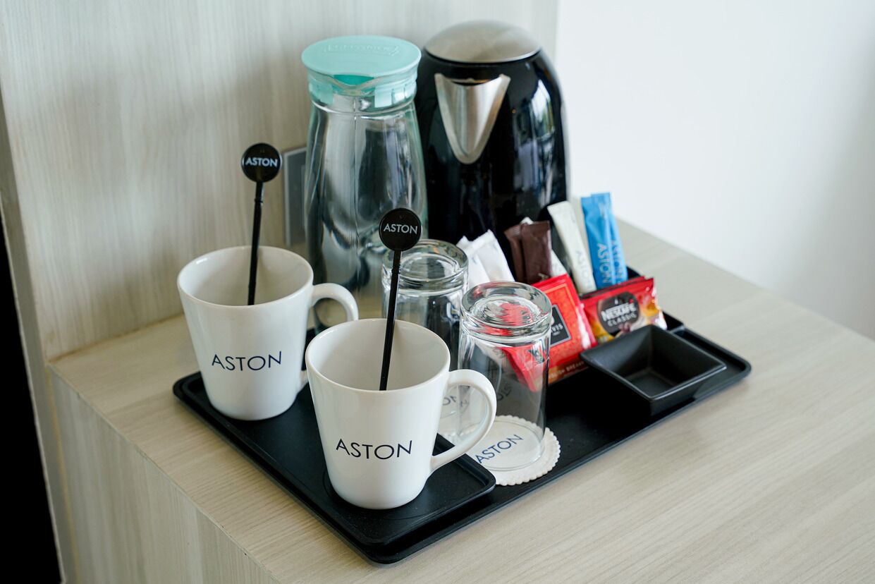 Two white cups with the word "ASTON" on a tray with a coffee maker, a glass, and some chocolate bars.