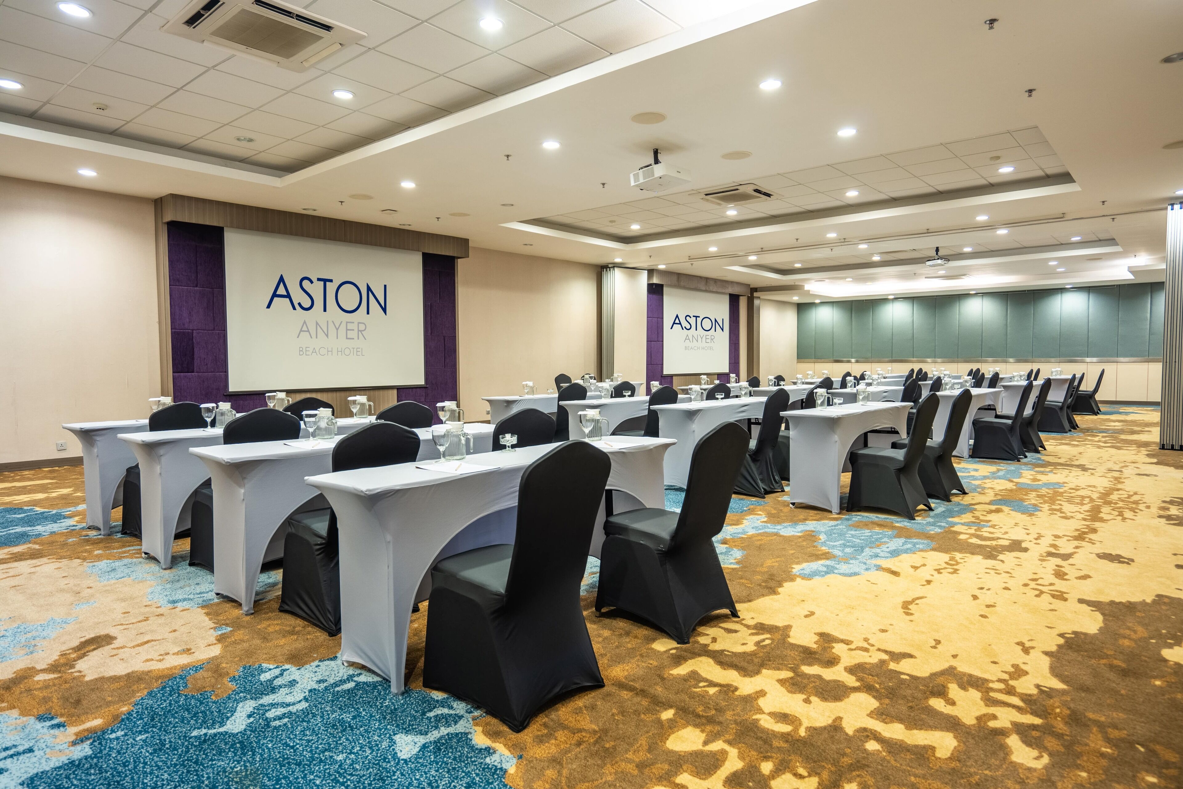 Un salón de conferencias en el Aston Anyer Beach Hotel con varias mesas, sillas y pantallas para presentaciones.