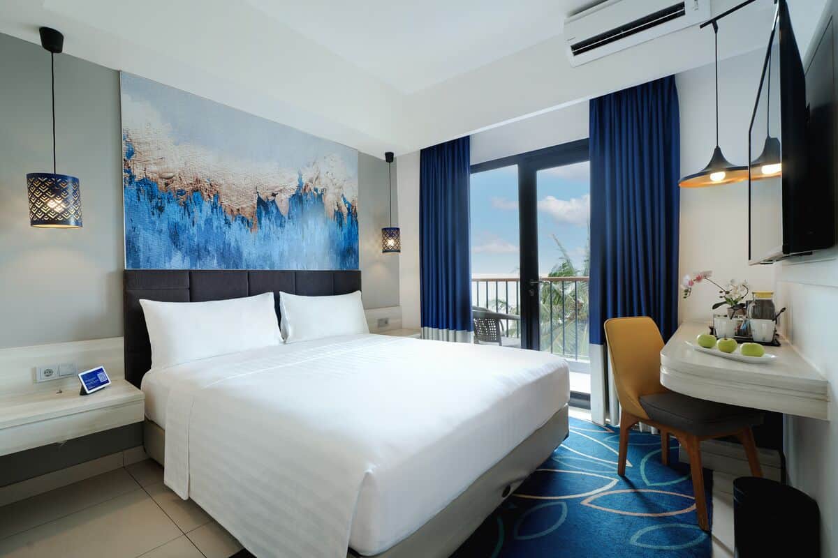Une chambre d’hôtel avec un grand lit, un bureau avec une chaise, un tableau au mur et un balcon avec des rideaux.