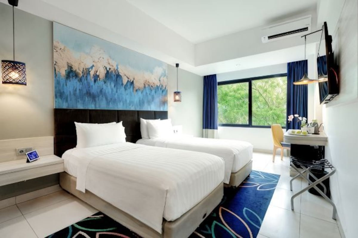 Habitación de hotel con dos camas, un escritorio, un televisor y un ventanal grande.