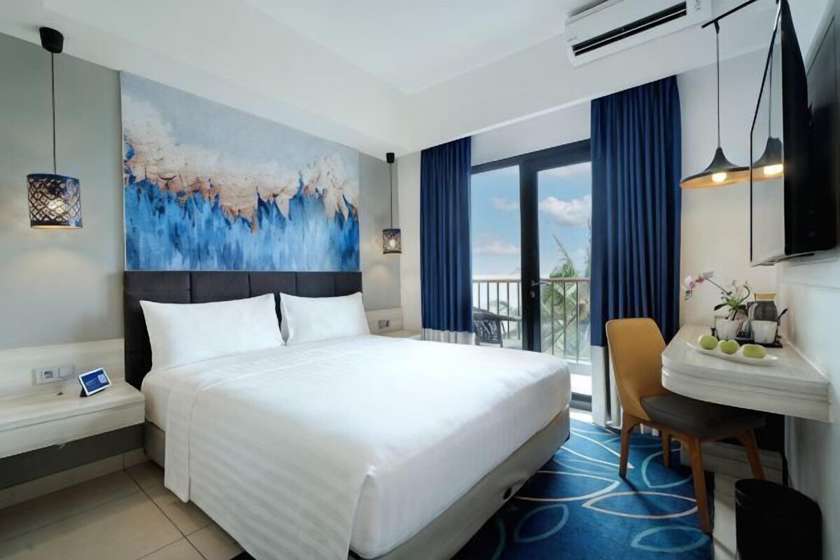 A hotel room with a large bed, a desk with a chair, a painting on the wall, and a balcony with curtains.