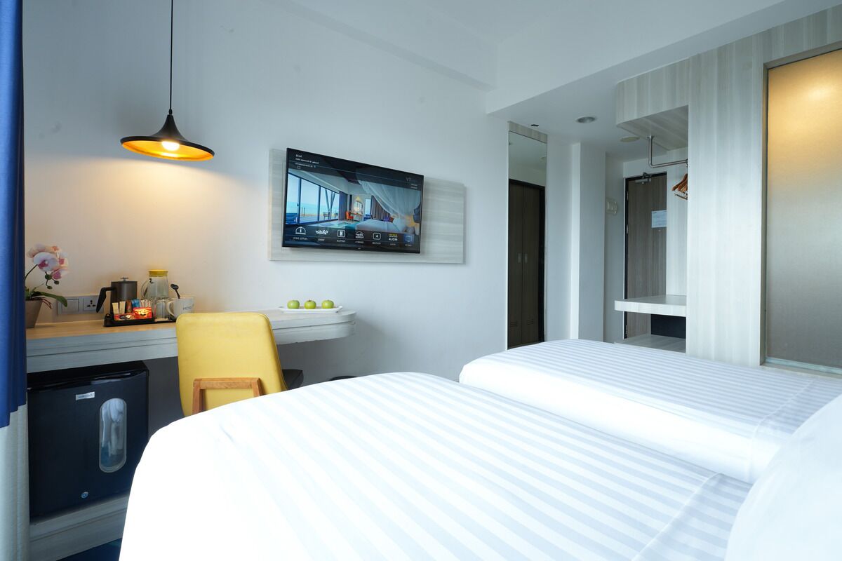 Ein Hotelzimmer mit zwei Betten, einem Schreibtisch mit Computer, einer kleinen Kühlschrank und einem an der Wand befestigten Fernseher.