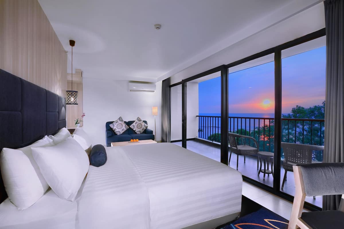 Une chambre d’hôtel moderne dotée d’un grand lit, d’un balcon avec mobilier d’extérieur et offrant une vue sur le coucher de soleil.
