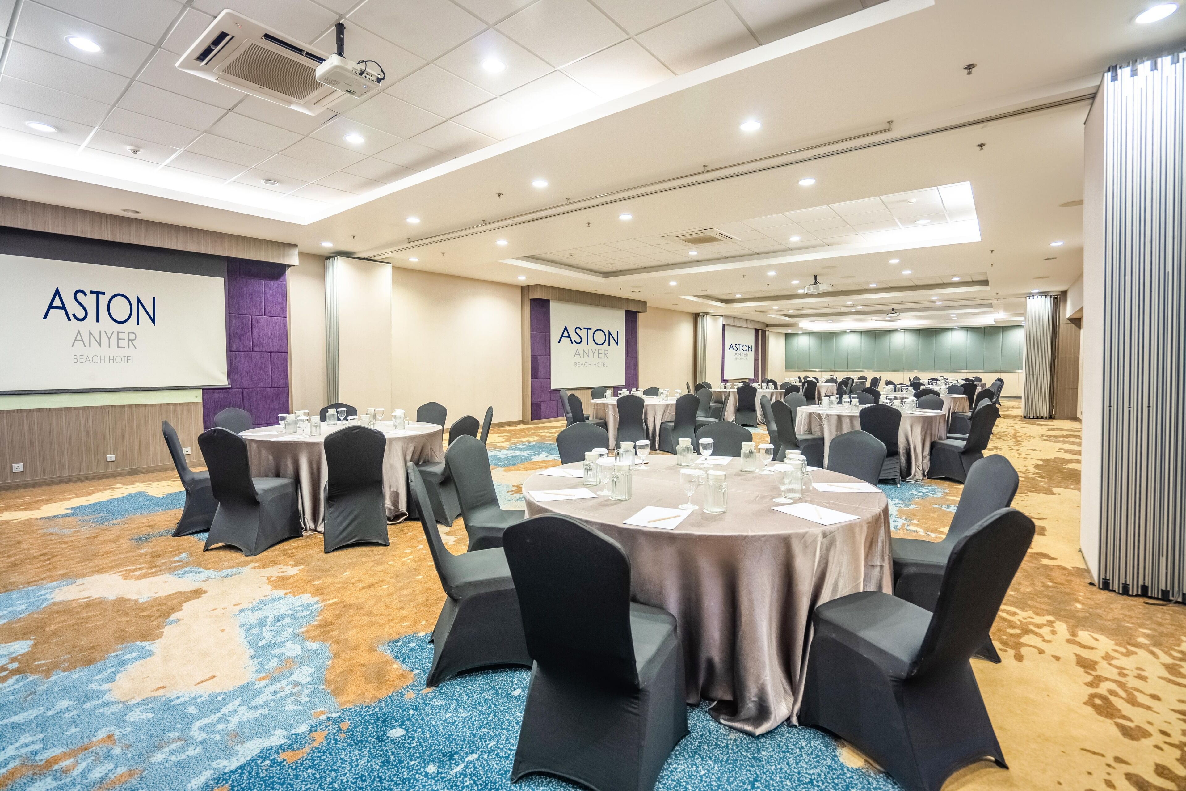 A conference room with round tables, black chairs, and a large screen displaying the hotel name.
