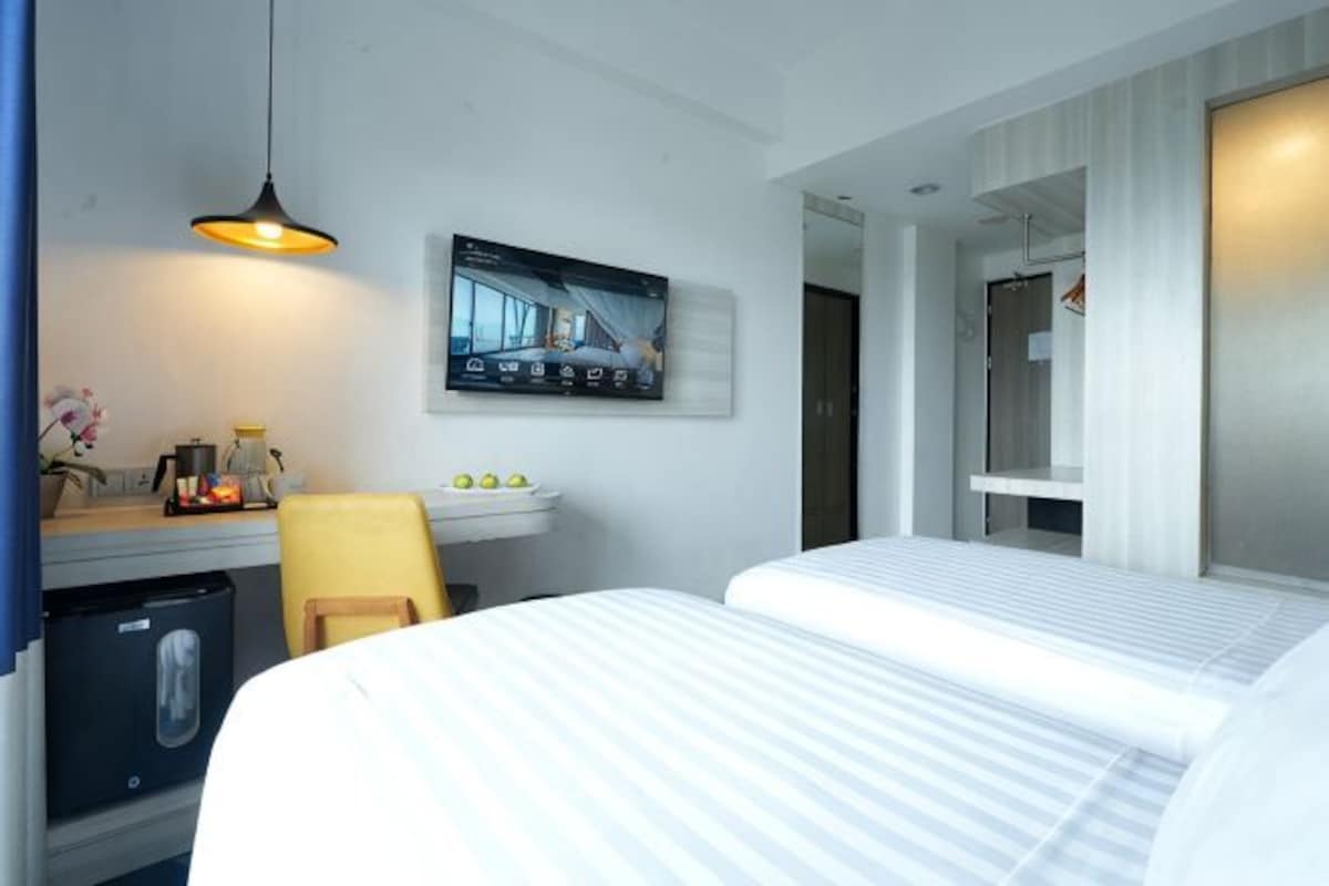 Habitación de hotel con dos camas, un escritorio con computadora, una silla, un televisor montado en la pared y la puerta del baño.