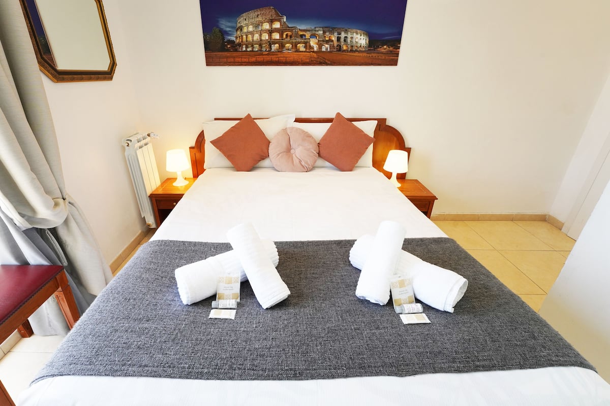 A neatly made bed with a grey blanket, two rolled towels, and a pillow. There are two bedside lamps, a framed picture of a building on the wall, and a window with curtains.