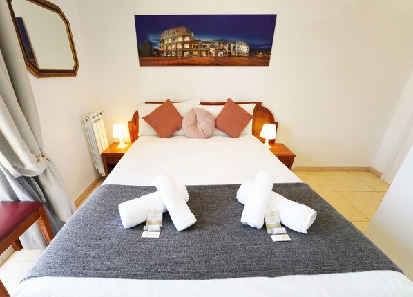 A neatly made bed with a grey blanket, two rolled towels, and a pillow. There are two bedside lamps, a framed picture of a building on the wall, and a window with curtains.