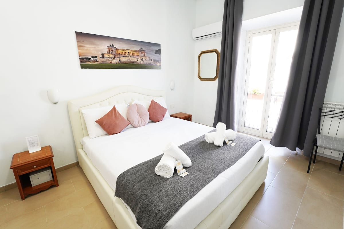 A hotel room with a bed, a nightstand, a bedside table, a towel, a window with curtains, and a wall-mounted air conditioner.