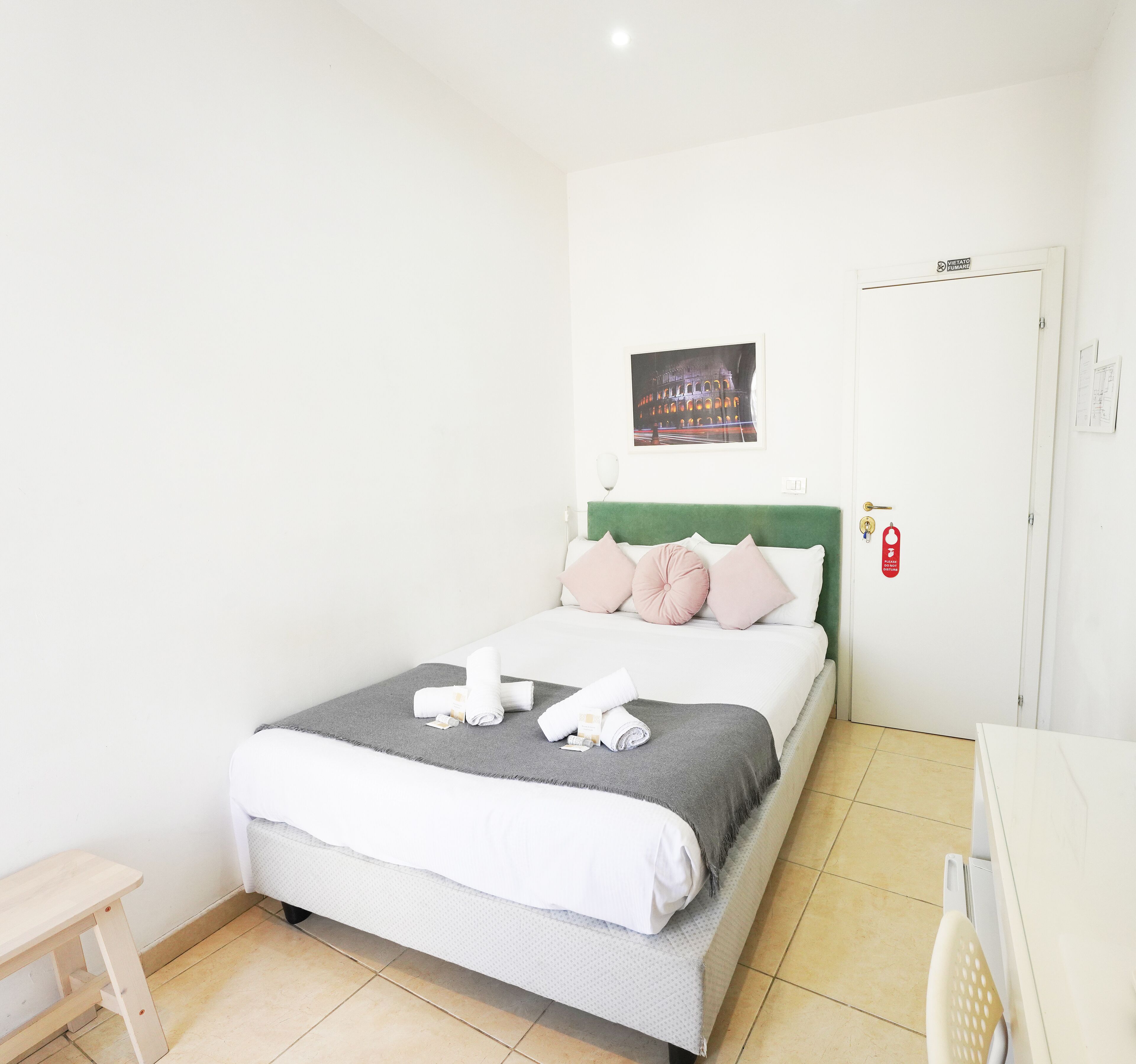 Cama bem arrumada com cabeceira verde, manta cinza e almofadas cor de rosa. Há um quadro de um edifício na parede e uma porta com uma placa vermelha.
