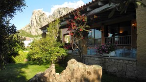 A rustic building with a stone facade, a balcony with potted plants, and a mountainous backdrop.
