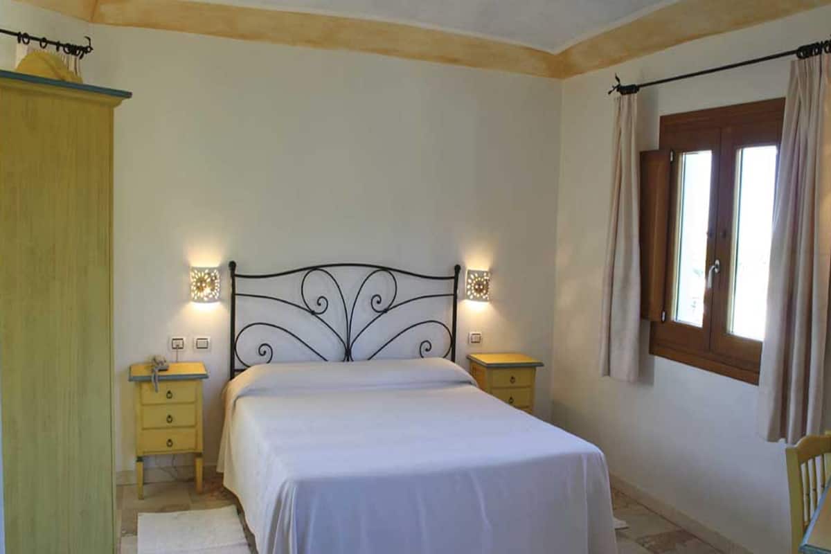 A bedroom with a bed, two bedside tables, a wardrobe, a window with curtains, and wall-mounted lights.