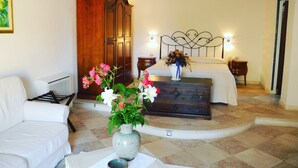 A hotel room with a bed, a wooden wardrobe, a white armchair, a vase with flowers, and a small table with a bowl.