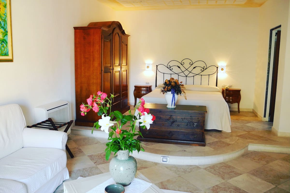 A hotel room with a bed, a wooden wardrobe, a white armchair, a vase with flowers, and a small table with a bowl.