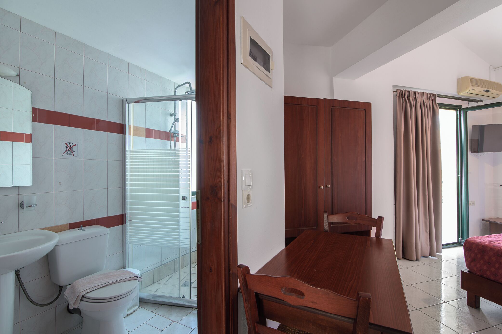 A bathroom with a toilet, sink, and shower. There is a wooden table with chairs and a bed with a red bedspread.