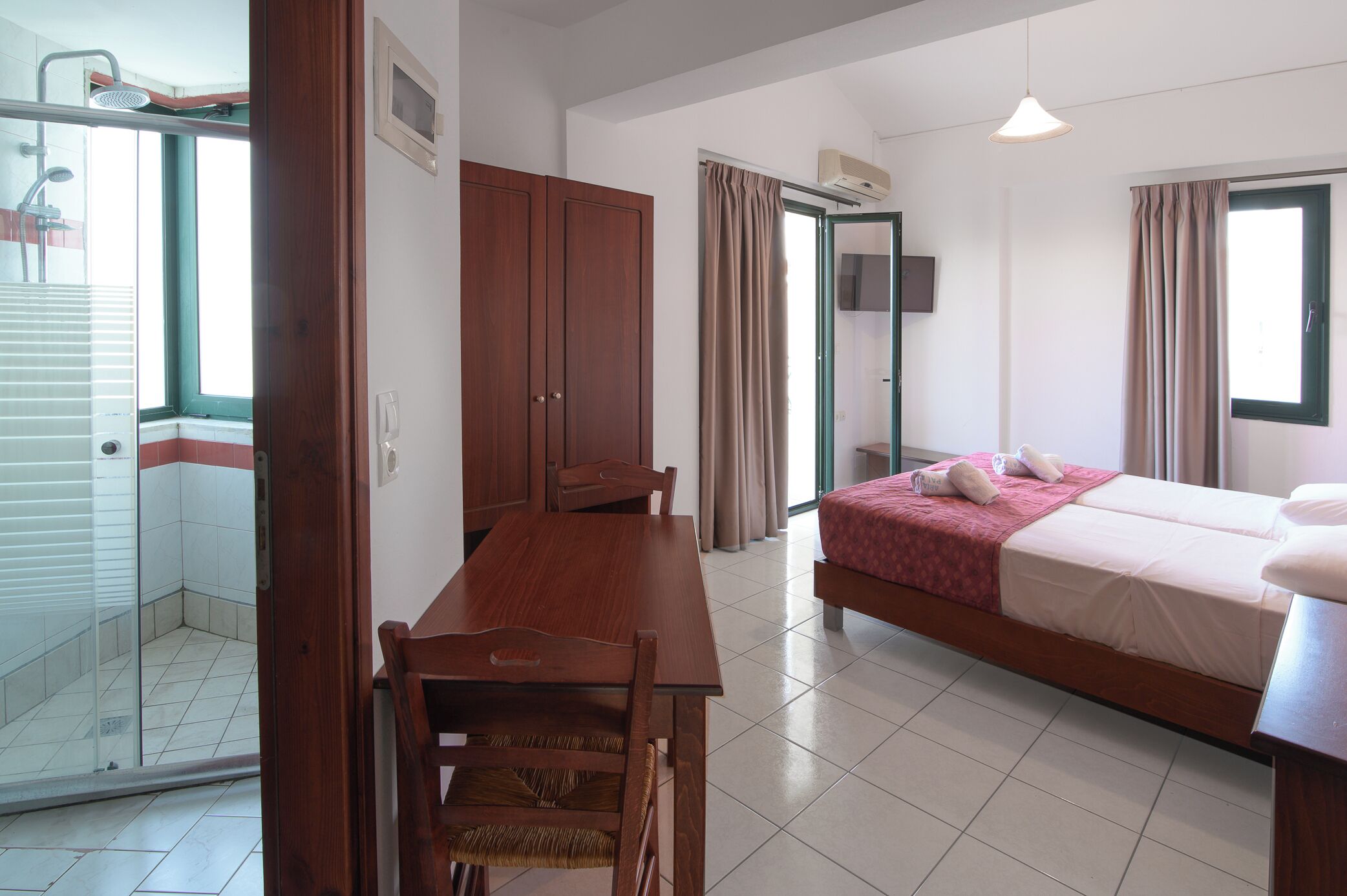 A hotel room with a bed, a wooden table, a chair, a shower area, and a window.