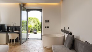 A modern living room with a grey sofa, a white chair, a large round table, and a glass door leading to an outdoor area with a view of greenery. - Ramo d'Aria Etna Boutique Hotel (Giarre)