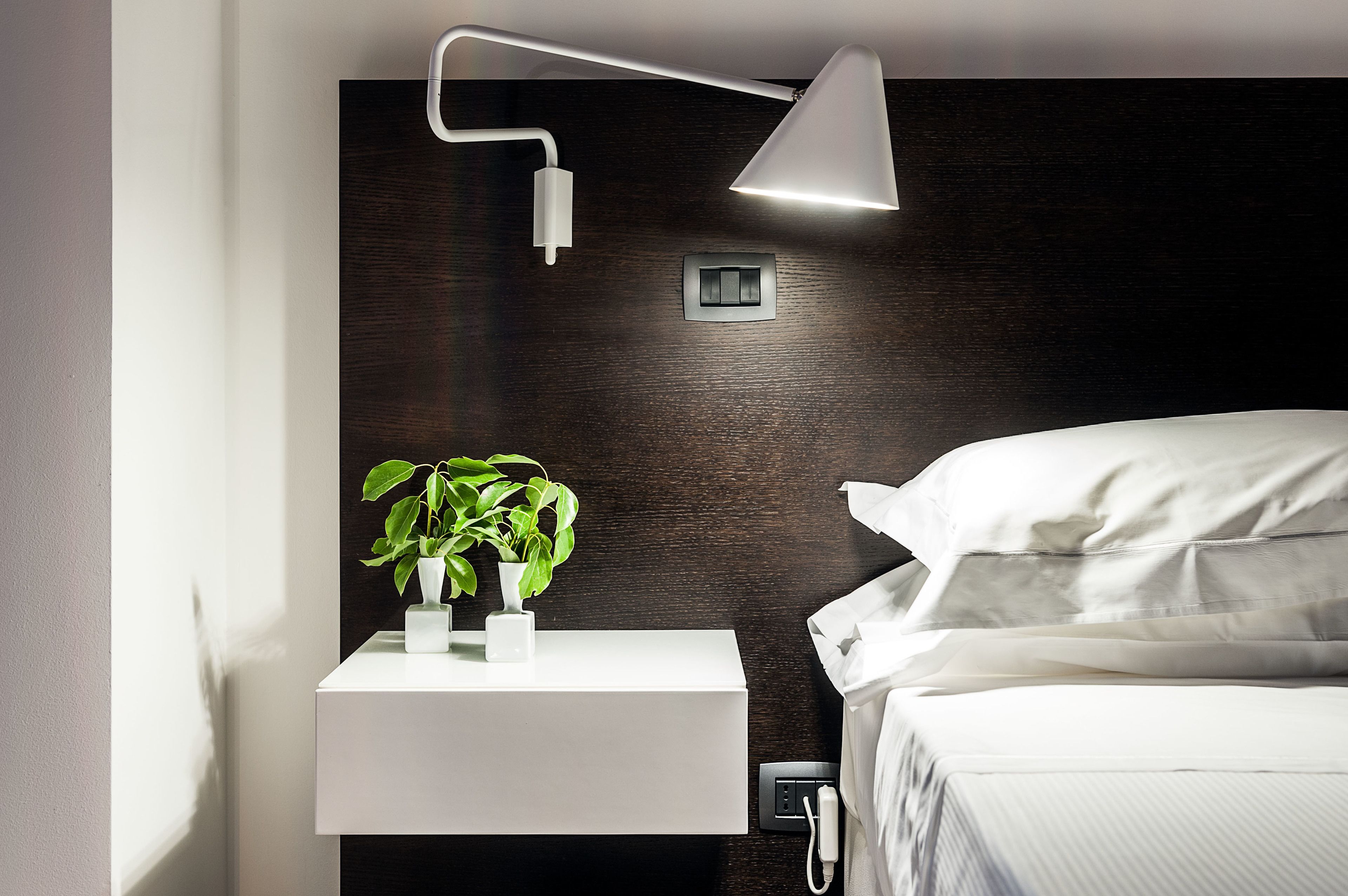 A modern bedroom with a white bedside table, a bedside lamp, a power outlet, and a potted plant.
