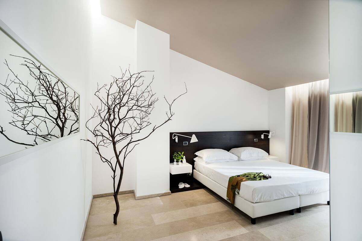 A modern bedroom with a large bed, a nightstand, a small table with a plant, and a tree decoration on the wall.