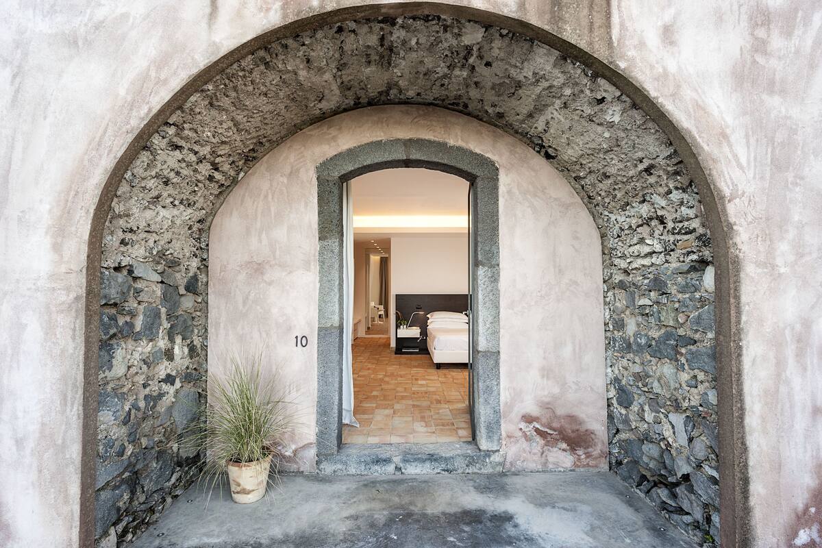 An arched doorway leading to a room with a bed, a potted plant, and a tiled floor.