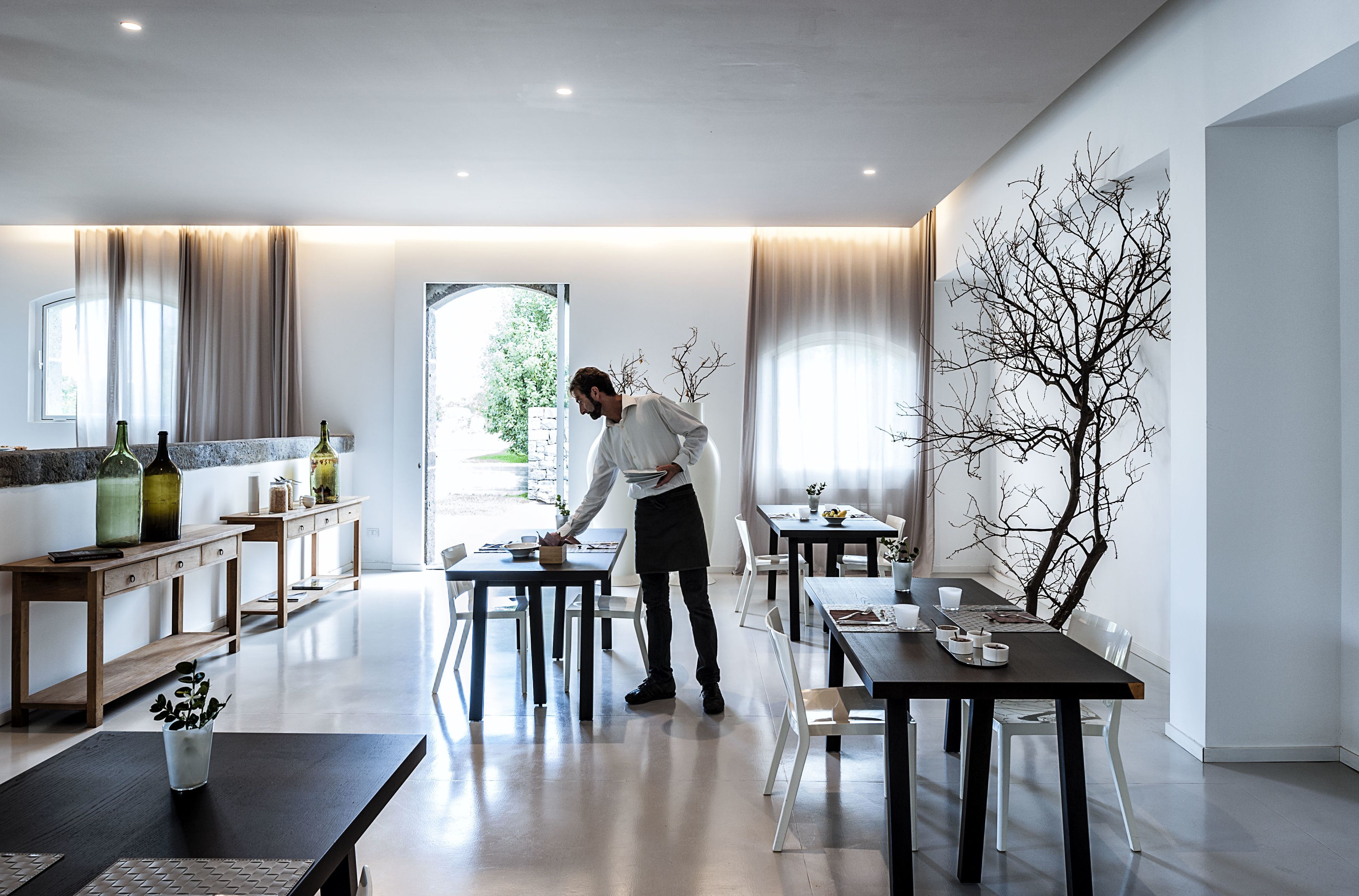 A modern dining area with tables and chairs, a waiter, and a large tree artwork on the wall.