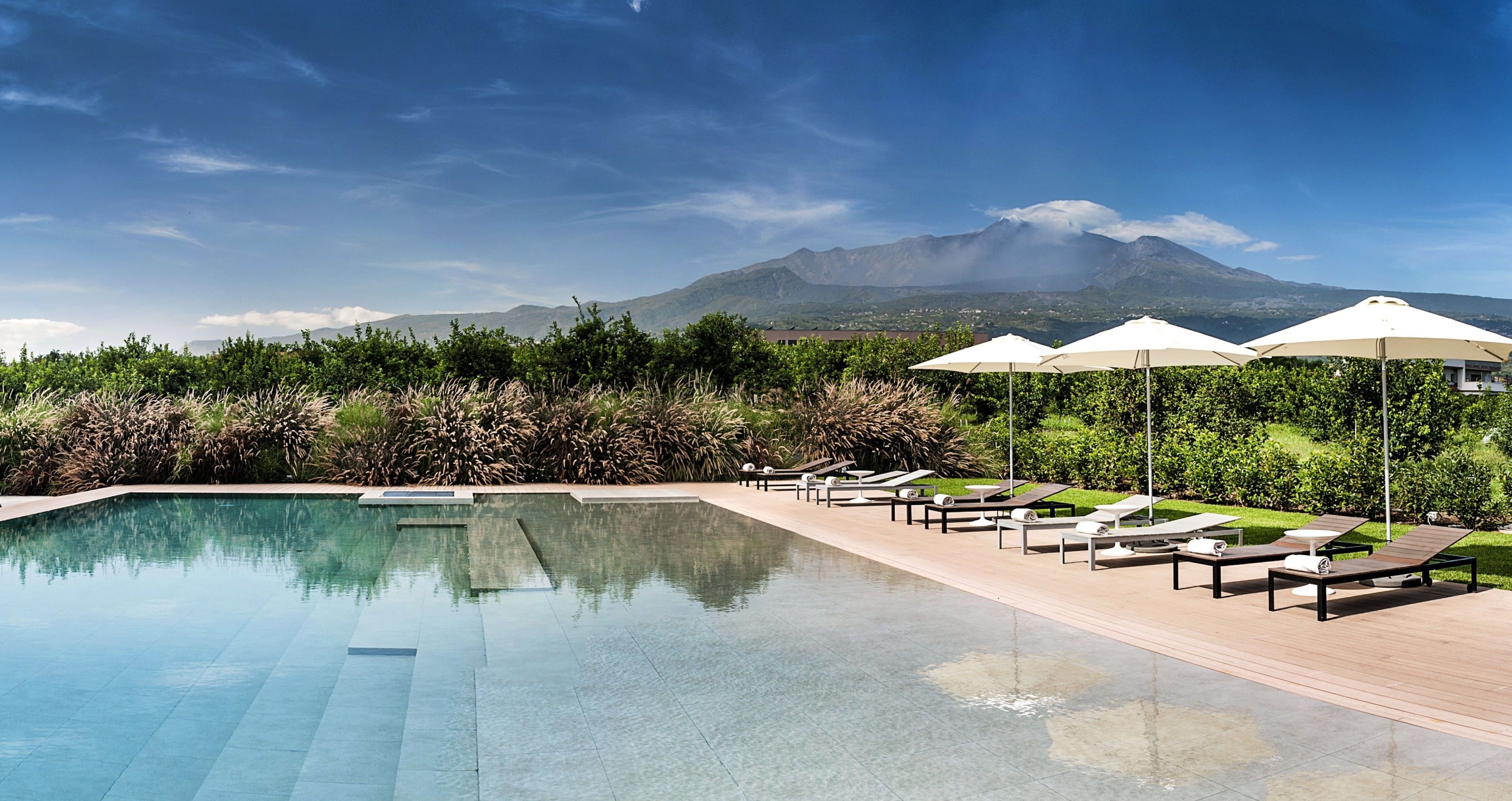 A swimming pool with lounge chairs and umbrellas, surrounded by greenery and a mountain view.