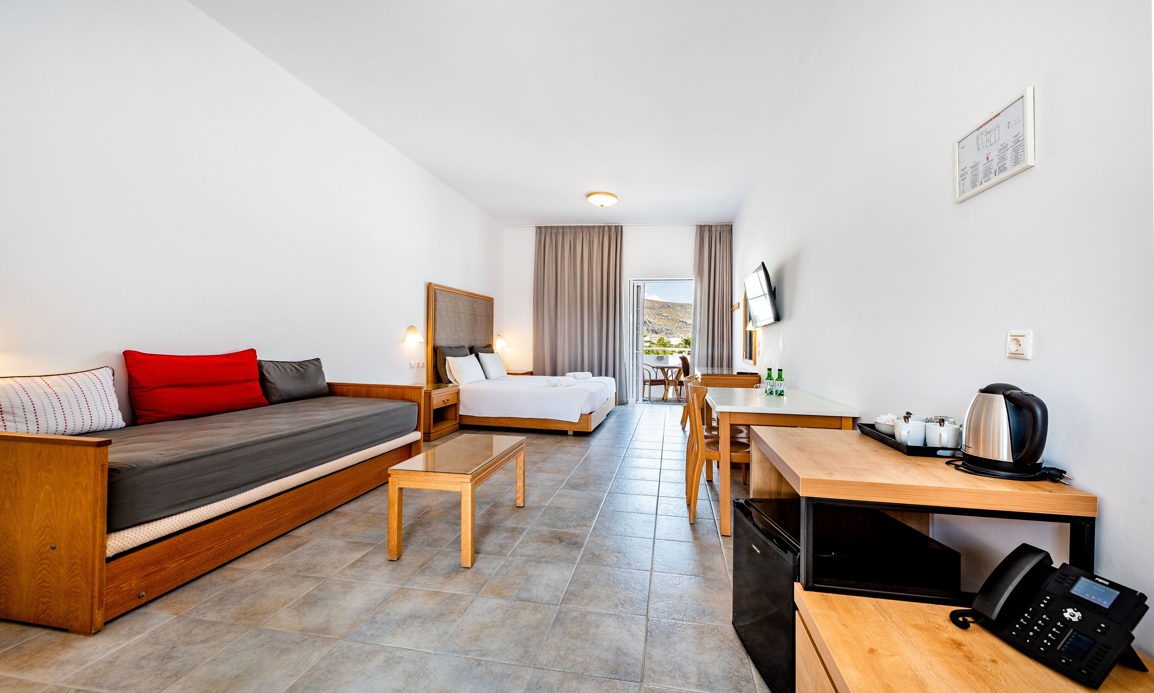 A modern hotel room with a bed, a sofa, a dining table, and a kitchen area.