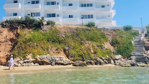 A white multi-story building with balconies, located on a hillside overlooking a beach. - Klinakis Beach Hotel (Chania)