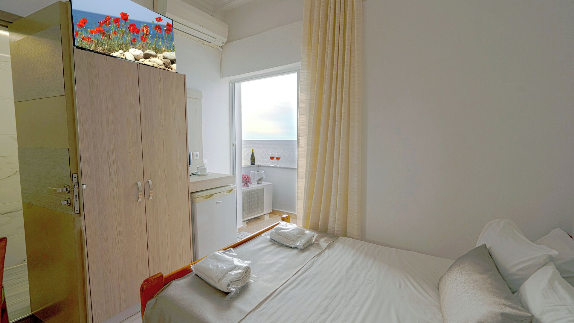 A hotel room with a bed, a wardrobe, a window with a view of the sea, and a small table with a bottle and glasses.
