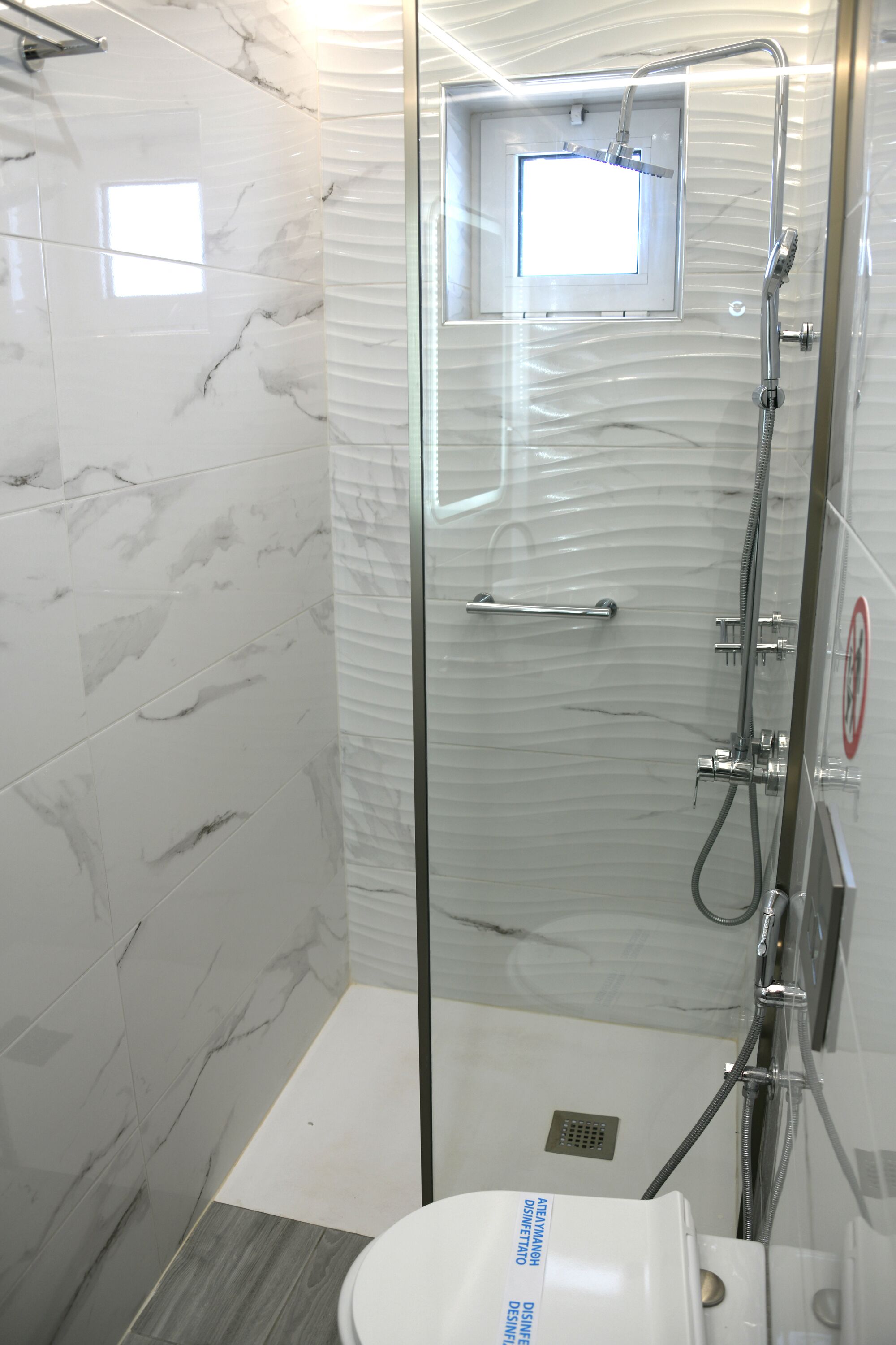 A modern bathroom with a glass-enclosed shower, marble tiles, and a small window.