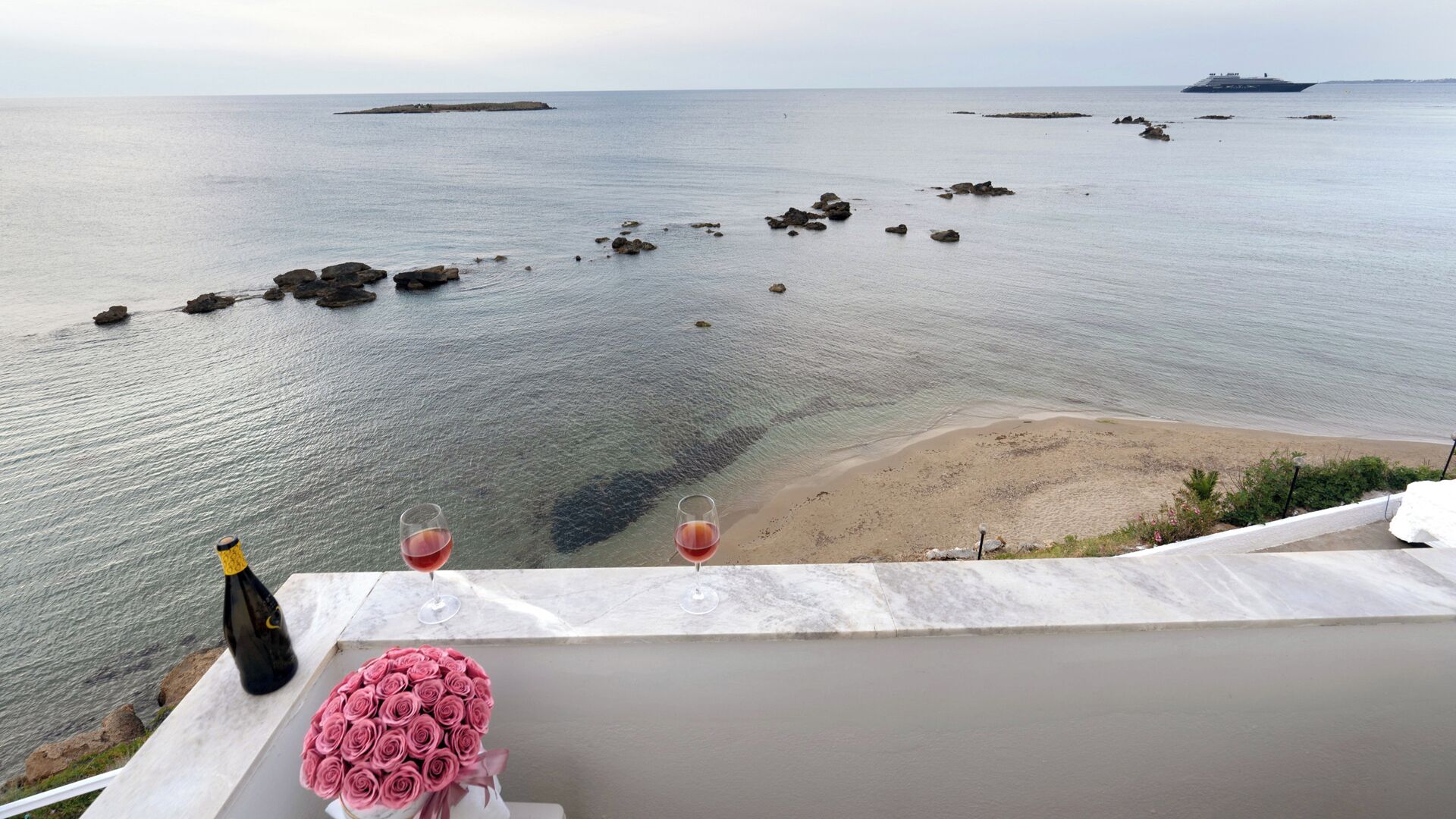 Vista para o mar com varanda, mostrando uma garrafa de vinho, dois copos de vinho e um buquê de rosas cor-de-rosa.
