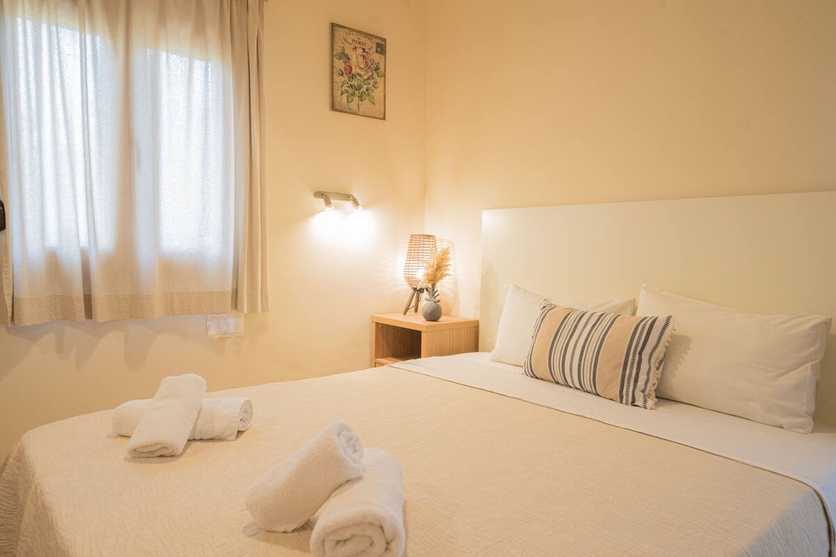 A neatly made bed with rolled towels, a bedside lamp, and a small table with a vase.