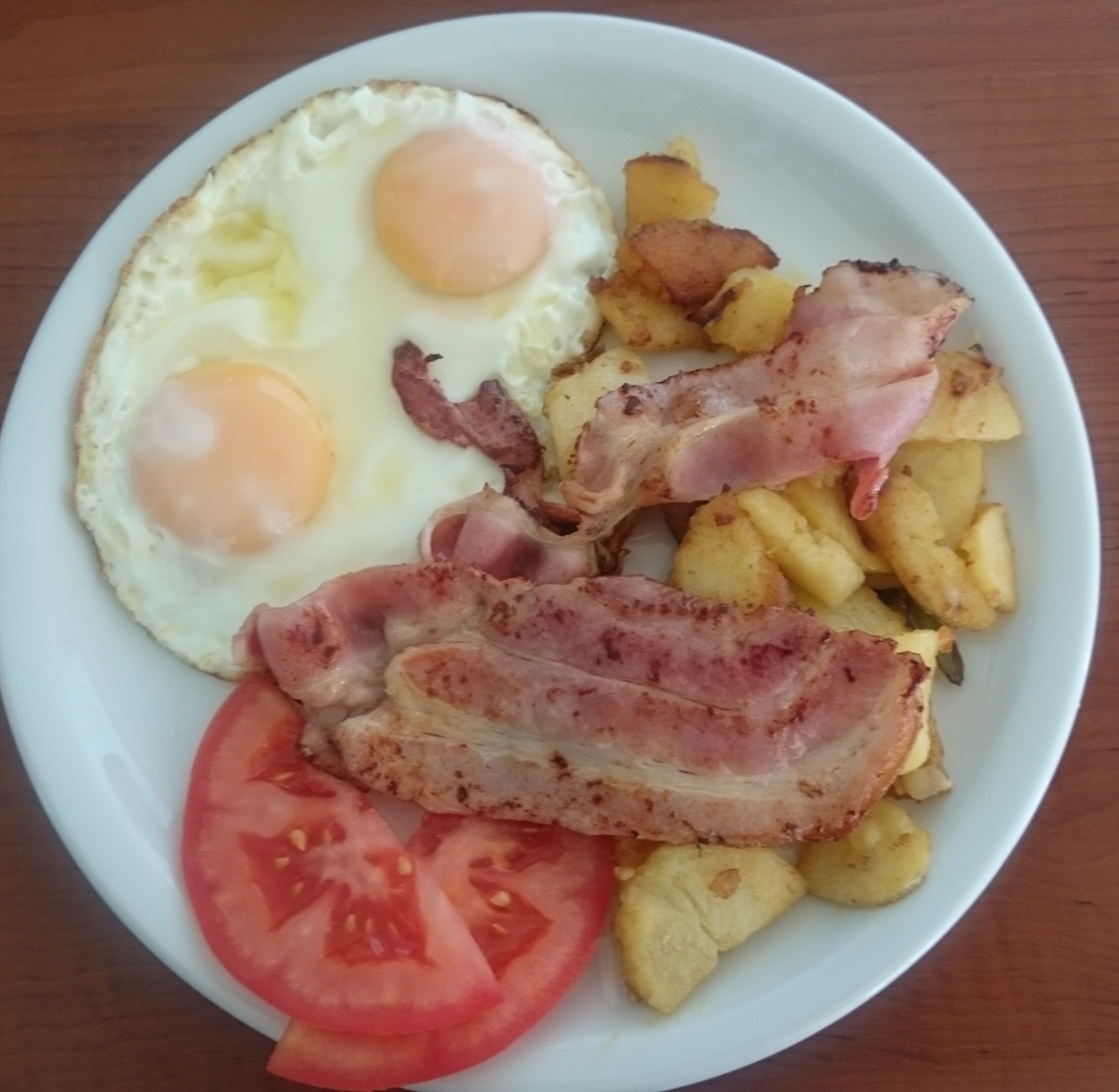 Une assiette avec deux œufs frits, des lardons, des pommes de terre rôties et une tranche de tomate.