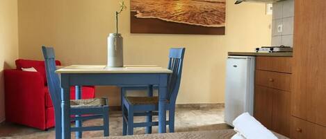 A room with a bed, a table with chairs, a small kitchenette, and a painting of a sunset over the ocean.
