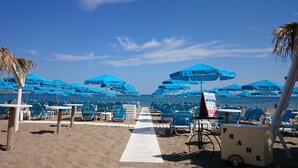 A beachside area with rows of blue umbrellas labeled "FRANK" and beach chairs arranged along a white walkway. - Frank Apartments (Malevizi)