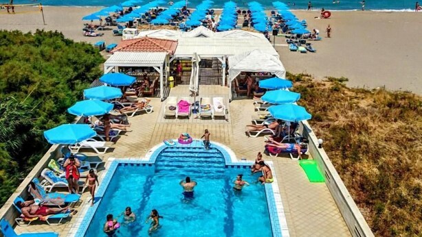 A pool area with lounge chairs and umbrellas, a beach in the background, and a building with a tiled roof. - Frank Apartments (Malevizi)