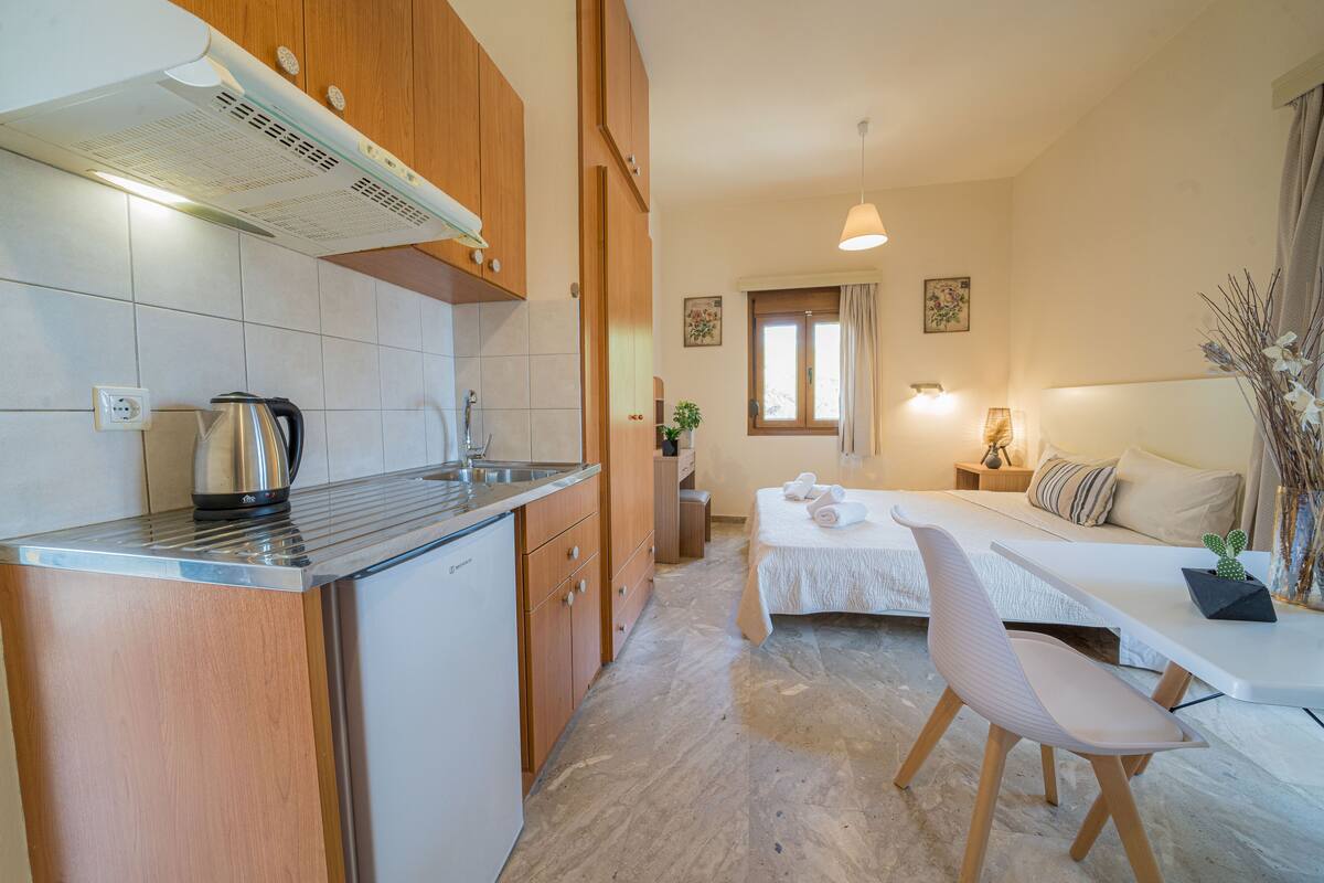 A compact kitchen with a kettle, a sink, and wooden cabinets. A bed with a white headboard and a bedside table with a lamp and a cactus is visible.