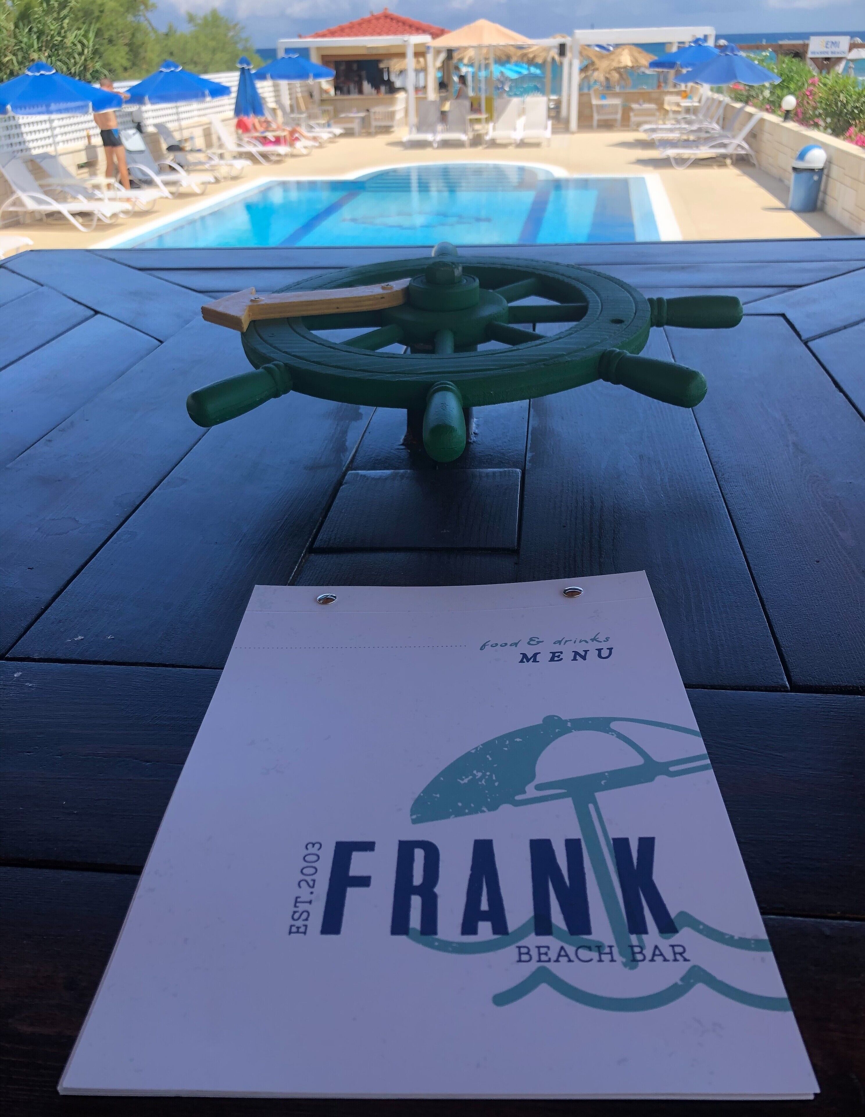Une table en bois avec un menu du Frank Beach Bar, un espace piscine avec des chaises longues et des parasols, et une grande roue verte.
