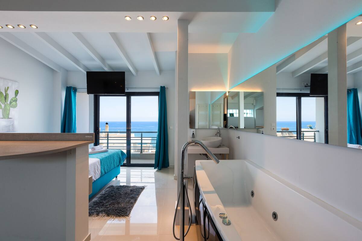 A modern hotel room with a balcony, a large mirror, a bathtub, and a view of the sea.