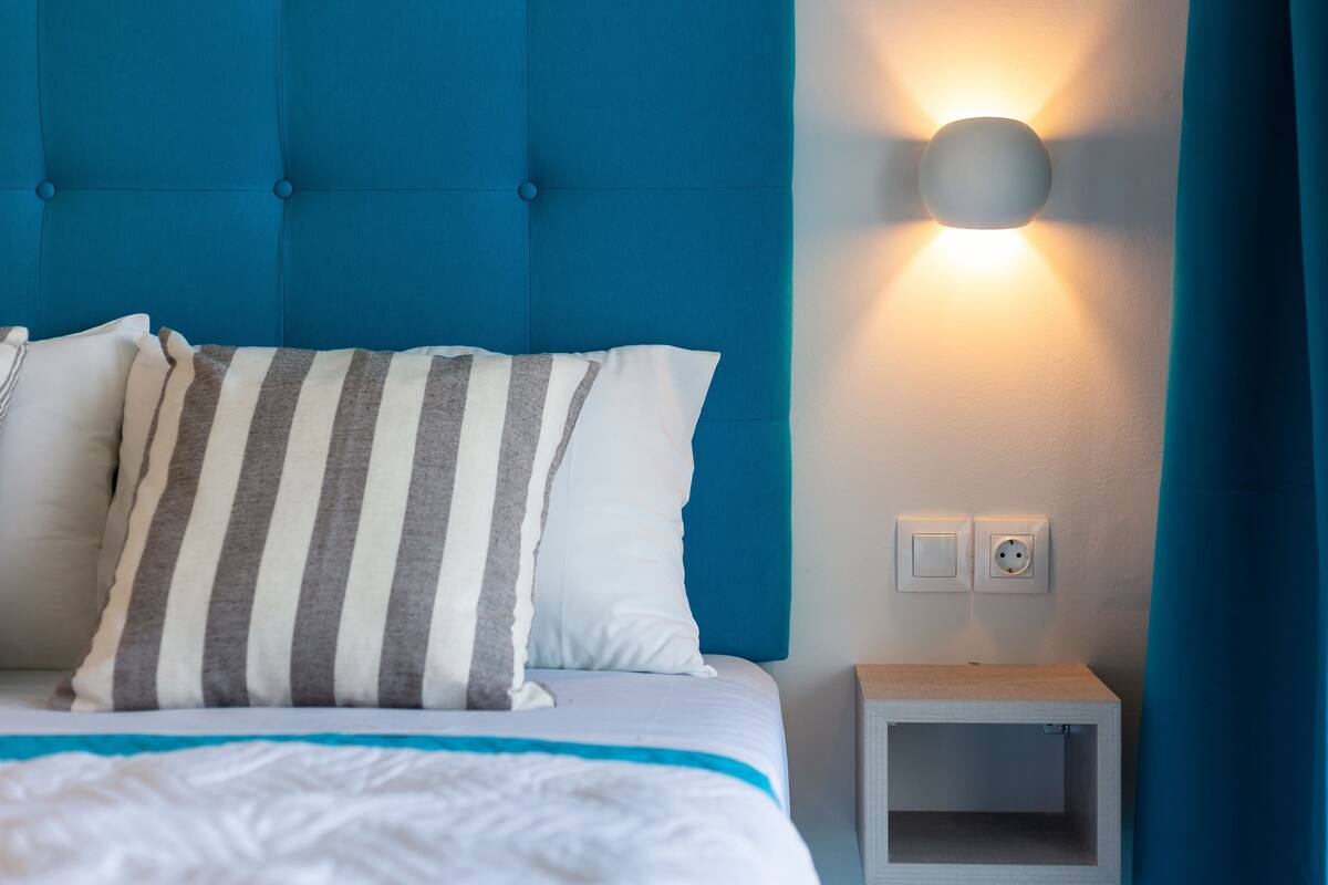 A bed with a blue headboard, white and striped pillows, and a bedside table with a power outlet.