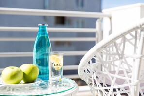 Une table en verre avec une bouteille, deux pommes et un verre d’eau sur un balcon.