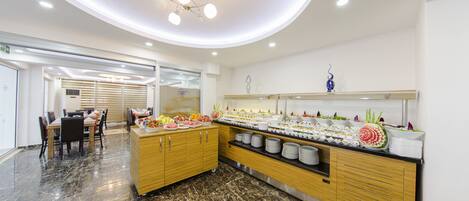 Um hall de hotel moderno com piso de mármore, área de buffet com diversos alimentos e um espaço de estar com mesas e cadeiras.