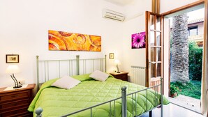 A bedroom with a green bed, two bedside tables with lamps, a radiator, and a large window with a view of greenery and a palm tree.