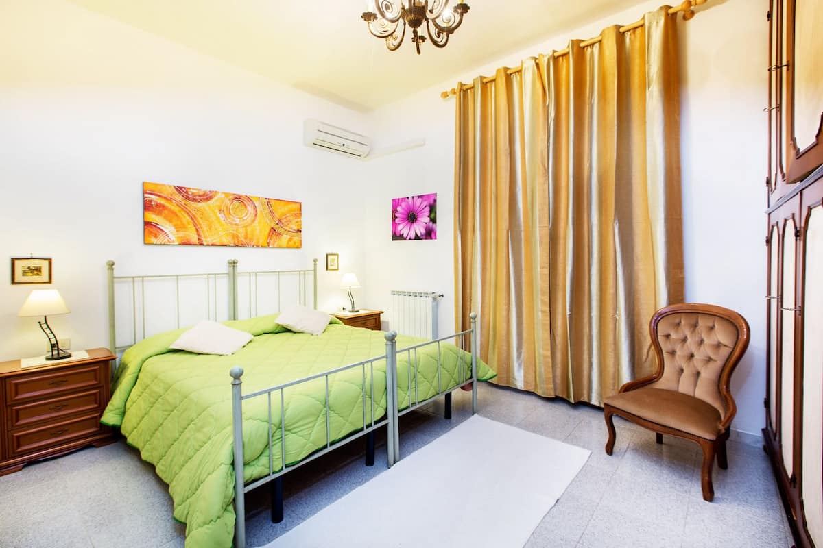 Um quarto com cama, cadeira, mesa de cabeceira, abajur, radiador e janela com cortinas.